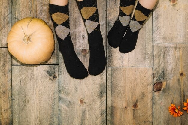 Two pairs of feet on wooden flooring wearing matching black, gold, and grey argyle socks next to a large pumpkin.