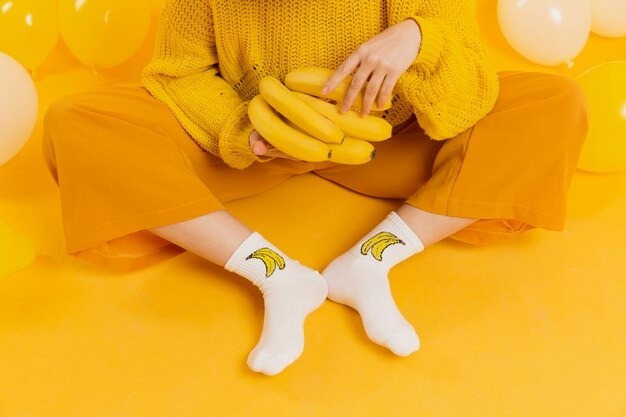 A person sitting cross-legged on a yellow background, wearing a yellow sweater, mustard pants, and white socks with a banana print while holding a bunch of bananas.