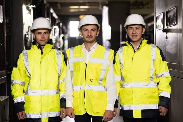 Three male industrial workers wearing white hard hats and yellow high-visibility safety jackets standing side-by-side.