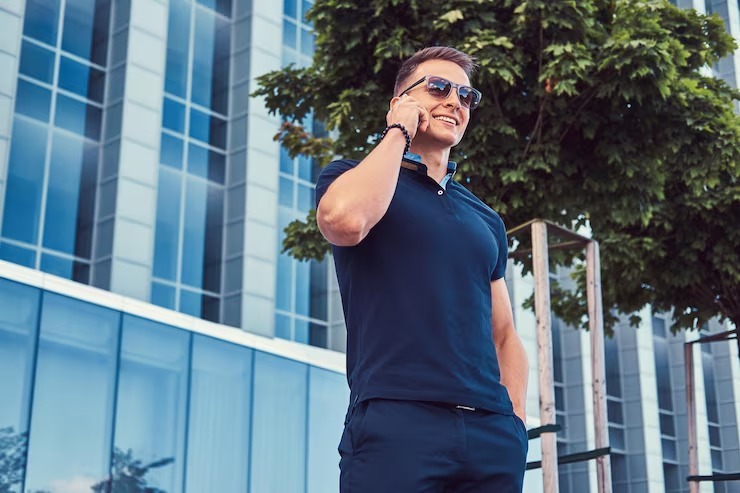 A smiling young man in a dark blue polo shirt and sunglasses talking on his phone outside a modern office building.